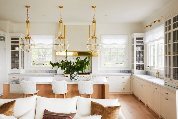 Bright, refined open-plan kitchen with extensive white shaker-style cabinetry, glass-front display cupboards, a central island with timber paneling and three upholstered stools, marble-look benchtops, and three prominent brass-and-glass pendant lights. A living area with a sofa is visible in the foreground, while large windows provide abundant natural light.