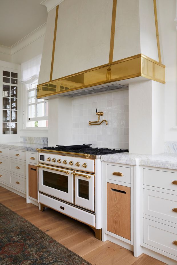 A bright, traditional-style kitchen featuring a large freestanding white range cooker with brass hardware, white paneled cabinetry, marble-look benchtops, a glossy white tiled splashback, and a substantial custom rangehood with brass trim. Glass-front cabinetry, tall windows, timber flooring, and a patterned rug complete the refined interior.