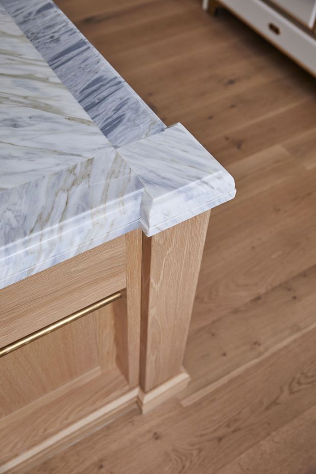 Close-up detail of a refined kitchen island or built-in joinery, featuring a veined white-and-grey marble benchtop with a waterfall-style edge, light timber cabinetry and support detailing, and a slim brass rail handle. Warm timber flooring and matching built-in furniture are visible in the background.