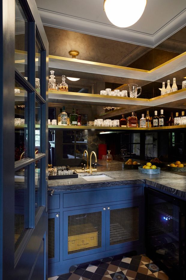 A compact, richly detailed home bar with dark blue built-in cabinetry, open illuminated glassware shelves, mirrored walls, liquor bottles, a stone bar counter, and brass-toned fixtures. The geometric black, white, and brown tiled floor and dark reflective finishes create a dramatic, luxurious atmosphere.