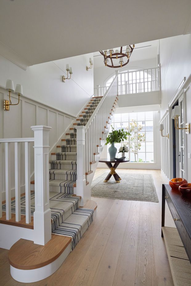 Bright, refined residential entry hall with a prominent staircase. The space features white painted wall panelling, a carpeted stair runner with geometric striping, white balustrades and decorative newel posts, pale timber flooring, brass-and-white wall sconces, and a large traditional chandelier. A console table, patterned rug, greenery, and large gridded windows create an airy, elegant foyer.