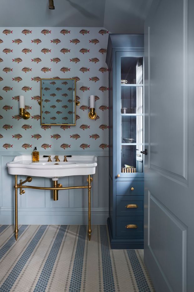 A compact, elegantly detailed bathroom viewed through an open paneled door. The room features a wall-mounted white porcelain basin with exposed brass plumbing, brass sconces and fittings, blue-gray painted wall panelling, fish-patterned wallpaper above the dado, a gold-framed mirror, a tall dark blue built-in cabinet with glass doors and drawers, and a patterned small-format tiled floor.