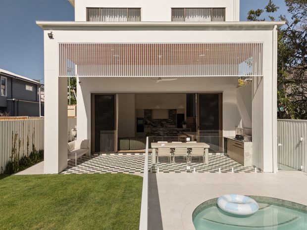 A contemporary white multi-level house viewed from the rear, featuring a covered outdoor entertaining area with a vertical slatted privacy screen, outdoor dining setting, built-in barbecue cabinetry, and a black-and-white checkered tiled terrace. A swimming pool occupies the foreground beside a paved area and lawn. The open interior behind the terrace includes a kitchen and living space.