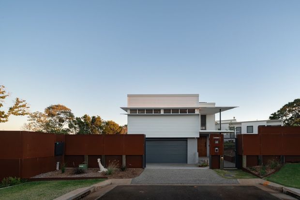 Street-facing view of a contemporary multi-level residence with a crisp white horizontal-clad facade, projecting flat roof forms, a dark sectional garage door, and a long rust-brown privacy fence. The driveway and pedestrian gate lead directly from the street, with landscaped planting along the frontage and neighbouring buildings visible behind.