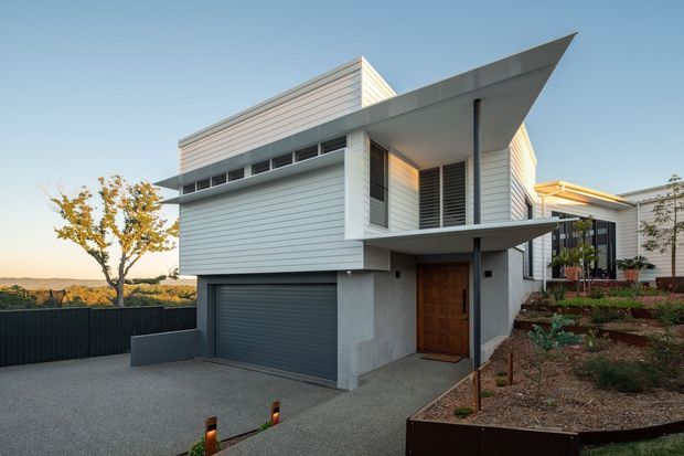 Exterior view of a contemporary multi-level residence with a sharply projecting asymmetrical roof canopy, white horizontal weatherboard cladding, louvred windows, concrete lower-level walls, a timber entry door, and an integrated dark garage door. The home sits on a landscaped sloping site with a driveway and retaining garden beds.