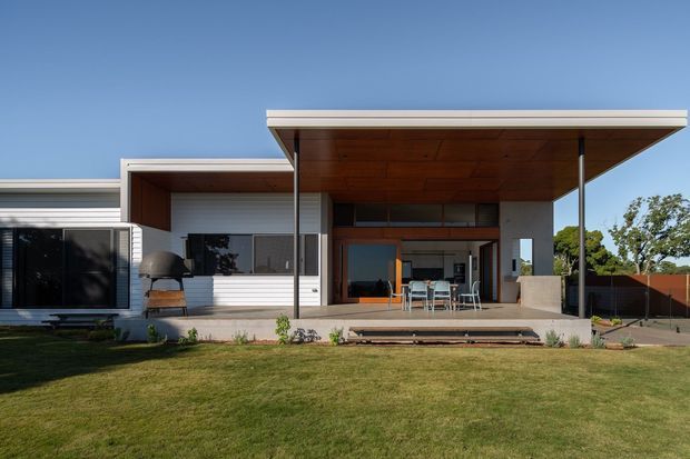 Single-storey contemporary house viewed from the rear garden, with a broad flat roof and deep covered outdoor living area. White horizontal cladding, warm timber soffit, large glazed doors, and a dining area create a strong indoor-outdoor connection. A manicured lawn and simple garden planting occupy the foreground.