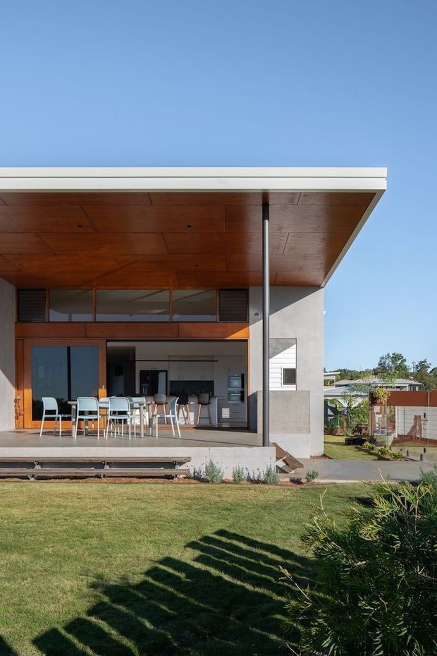 Exterior view of a modern single-storey home with a broad flat roof and deep covered patio. The patio ceiling and upper facade are clad in warm timber, with large sliding glass doors opening to a kitchen and dining area furnished with an outdoor table and chairs. The building combines white rendered walls, timber-framed glazing, a slender steel support post, and a raised concrete terrace overlooking a landscaped suburban lawn.
