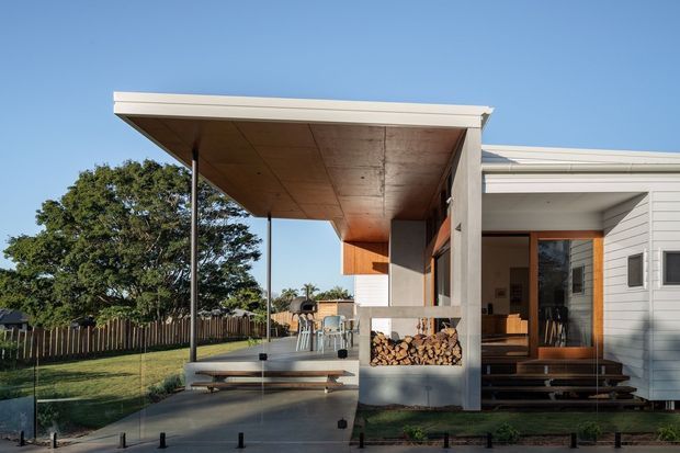 Exterior view of a contemporary single-storey home with a broad flat-roofed covered outdoor terrace, timber-lined soffit, concrete posts, and an open entry porch. The terrace includes built-in bench seating, an outdoor table, and neatly stacked firewood integrated beneath the structure. The adjacent facade uses light horizontal weatherboard cladding, with timber steps leading into the house and a fenced lawn beyond.