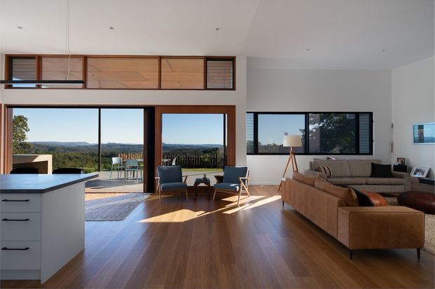 A bright open-plan living room with an integrated kitchen, extensive floor-to-ceiling glazing, timber-framed windows and doors, and views across a rural landscape. The room features warm timber flooring, a white kitchen island, a large tan sectional sofa, mid-century-inspired lounge chairs, and built-in timber-framed clerestory windows. Natural daylight creates a warm, airy atmosphere.