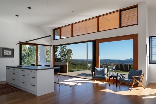 Bright open-plan interior combining a contemporary kitchen and living area, with a large white island, extensive timber-framed glazing, high clerestory windows, and views toward a wooded, hilly landscape. Warm timber flooring and mid-century-inspired lounge chairs create a refined, airy atmosphere.