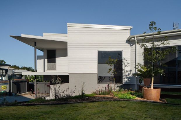 Exterior view of a contemporary low-rise home with a layered, flat-roof composition. The facade combines horizontal light-coloured weatherboard cladding with smooth grey rendered wall sections, dark-framed windows, broad roof overhangs, and a covered outdoor area. A landscaped lawn and garden sit in the foreground.