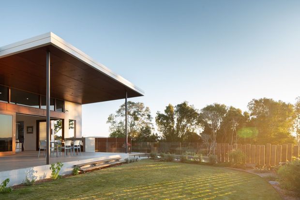 Modern single-storey pavilion-style home with broad flat roof overhangs, slender dark structural posts, extensive glazing, and a warm timber-lined soffit. The house opens onto a raised timber deck and landscaped lawn, with a simple outdoor dining area and distant tree screening. Warm late-afternoon sunlight creates a relaxed connection between the interior and garden.