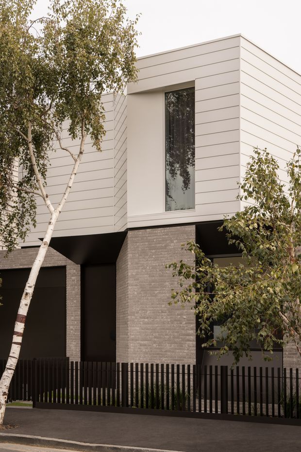 A contemporary multi-storey residential facade with a boxy, flat-roofed form. The upper level is clad in light horizontal weatherboard-style panels with a tall recessed window, while the lower level uses textured grey brick and dark recessed openings. A black vertical-slat fence and mature trees frame the street-facing elevation.
