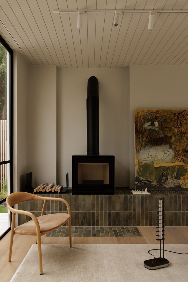 Warm, refined living room interior centred on a black freestanding wood-burning stove with a tall flue, set against a tiled hearth and feature wall. The room has a light painted timber-lined ceiling, large glazed opening, neutral walls, a sculptural timber lounge chair, soft rug, artwork, and track lighting. The hearth is finished in vertically arranged dark olive-grey rectangular tiles with lighter tiled accents on the floor.