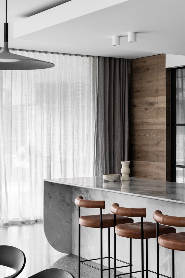 A refined contemporary kitchen or entertaining space with a curved grey stone island, three brown leather bar stools, sheer and dark layered curtains, a warm timber wall panel, and minimalist black-and-white lighting. The space has a restrained, luxurious material palette and soft natural light.