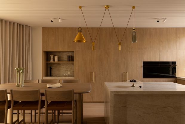 Warm, refined contemporary kitchen and dining area with extensive timber cabinetry, a stone island, integrated black appliances, a timber dining table, and pendant lighting. Vertical curtains and a linear timber ceiling add texture and softness.