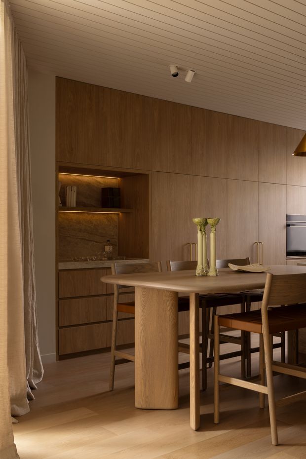 A warm, refined dining area with a round light-oak dining table and matching timber chairs, set against a full-height dark timber built-in wall. Integrated shelving and cabinetry feature concealed warm lighting, with a built-in appliance visible at right. The room has a slatted white ceiling, timber flooring, soft curtains, and understated decorative objects.