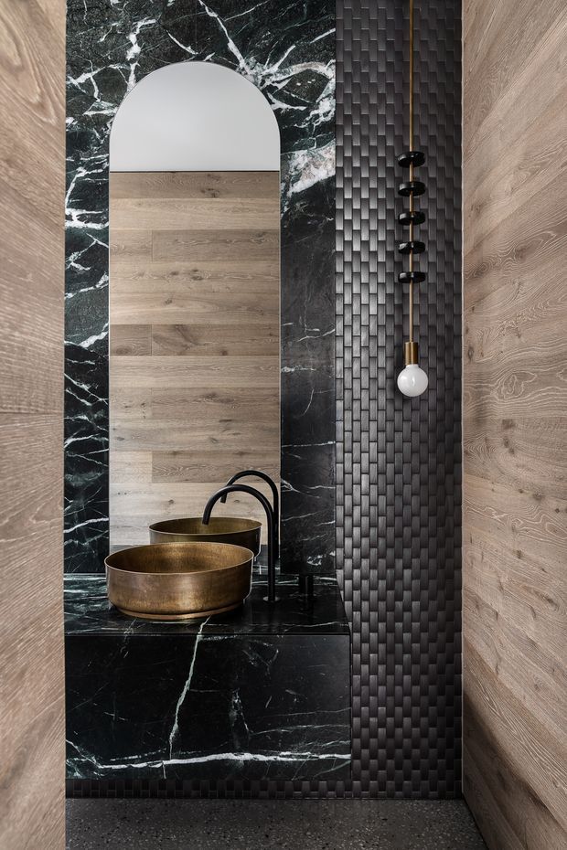 A dramatic, refined bathroom vanity alcove with twin bronze vessel basins and matte black curved faucets set on a black marble benchtop and vanity surround. A large arched mirror is framed by pale timber wall panelling and veined dark marble, while a vertical feature wall of dark textured rectangular tiles carries a hanging globe pendant. The floor appears dark speckled stone or terrazzo, and the overall composition is rich, highly contrasted, and luxurious.