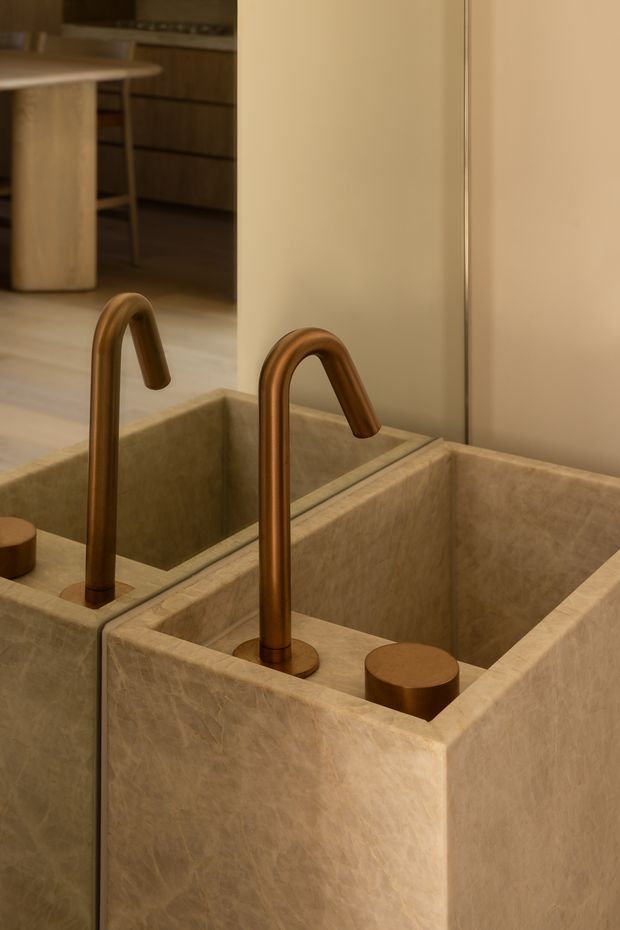 Close-up of a sculptural bathroom basin with a deep rectangular stone or stone-look vessel, paired with a tall curved copper-toned tap and cylindrical matching control. A large mirror reflects the basin and tap, while a warm timber-toned kitchen or living area is visible beyond. The image has a warm, refined, material-focused contemporary feel.