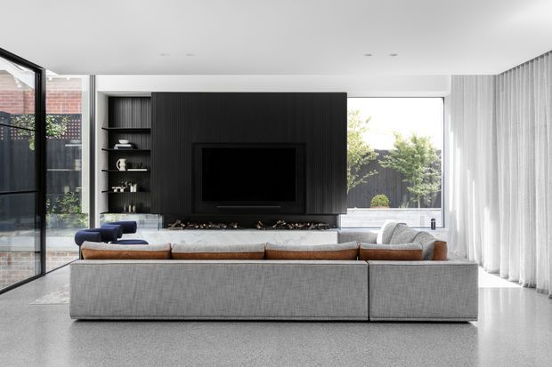 A bright contemporary living room viewed from behind a large modular sectional sofa. The room features a dark vertically ribbed media-wall partition with an inset television and open shelving, expansive glazing to a courtyard, sheer full-height curtains, a polished light terrazzo floor, and restrained neutral furnishings with warm timber and blue accents.