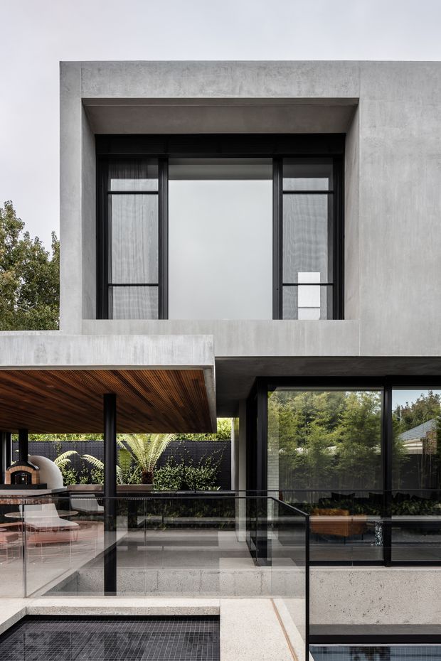 A contemporary multi-level residential exterior with a strong geometric concrete facade, large black-framed glazing, and an elevated terrace overlooking a partially visible tiled pool. The upper level features a deeply recessed window opening with sheer curtains, while the lower terrace includes a timber-lined roof or soffit, glass balustrades, planted screening, and an outdoor entertaining area. The composition is monochromatic and refined, with concrete, dark metal framing, timber, and abundant greenery.