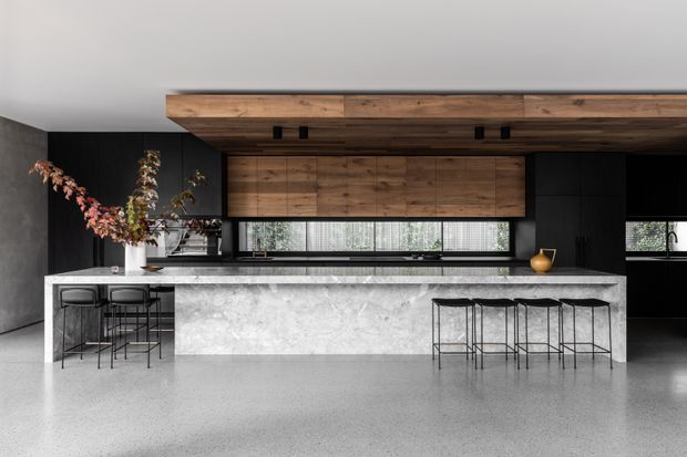 A spacious contemporary kitchen dominated by a long stone island with seating on both sides. The room features dark cabinetry and walls, extensive warm timber overhead cabinetry and a timber-clad ceiling beam, narrow horizontal windows, black bar stools, and a polished grey floor. The restrained black, grey, and timber palette creates a refined, dramatic atmosphere.