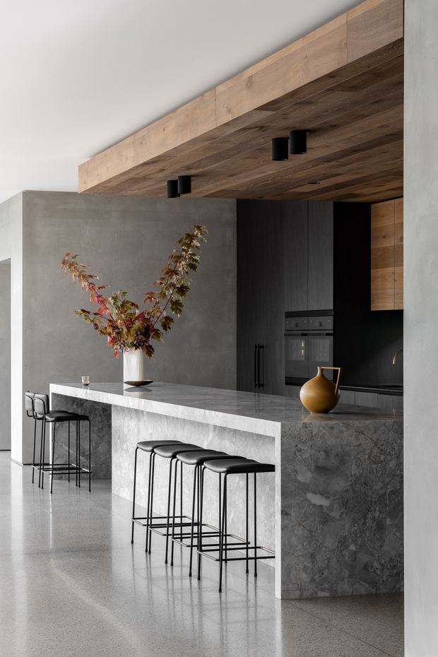 A sophisticated contemporary kitchen with a large marble island and integrated seating, dark timber and black cabinetry, built-in appliances, and a broad timber-lined ceiling soffit. Grey plaster walls and polished concrete or terrazzo flooring create a restrained, high-contrast palette, while sculptural ceramic decor and foliage add warmth.
