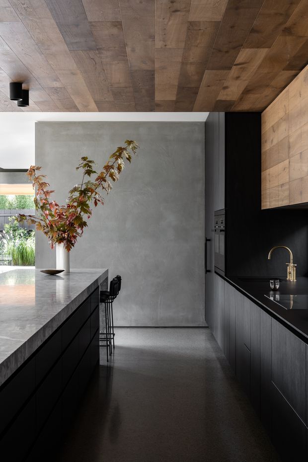 A refined contemporary kitchen with a long central island and dark, handleless cabinetry. The island has a pale stone benchtop and integrated seating, while the perimeter cabinetry combines black lower units, a black splashback, and warm timber overhead cupboards. A timber-panelled ceiling adds visual warmth, contrasted with a smooth grey concrete-look wall and dark polished flooring. A large opening brings in natural light and views of greenery.
