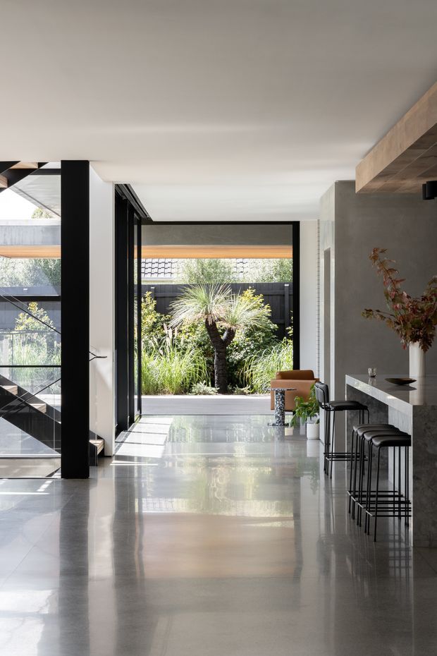 A bright, contemporary open-plan interior viewed along a polished concrete-finished circulation space. Full-height black-framed glazing and an open stairwell connect the interior to a landscaped courtyard. On the right, a long stone-topped bar or kitchen counter is paired with slim black bar stools, while a timber-lined overhead feature adds warmth. The palette is predominantly white, grey, black, and natural wood, with strong reflections and abundant daylight.