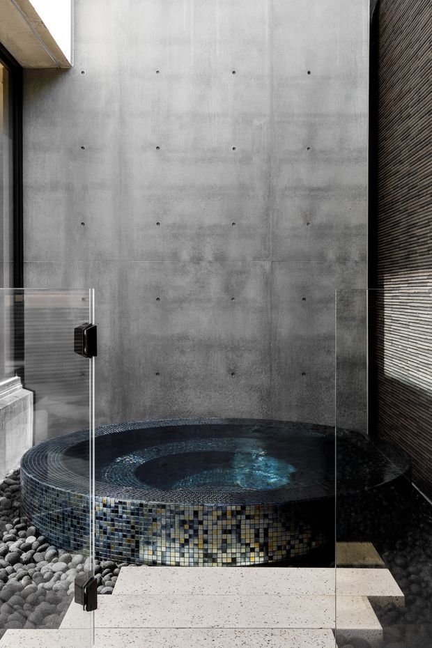 A dramatic contemporary spa bathroom featuring a round sunken or freestanding mosaic-tiled soaking tub with bubbling water, enclosed by clear frameless glass panels. The space has a raw grey concrete feature wall with visible formwork fixing points, a dark textured vertical wall surface, pebble flooring, and pale stone or concrete stepping slabs. The palette is dark, monochromatic, and tactile, with natural stone and water creating a luxurious, serene atmosphere.