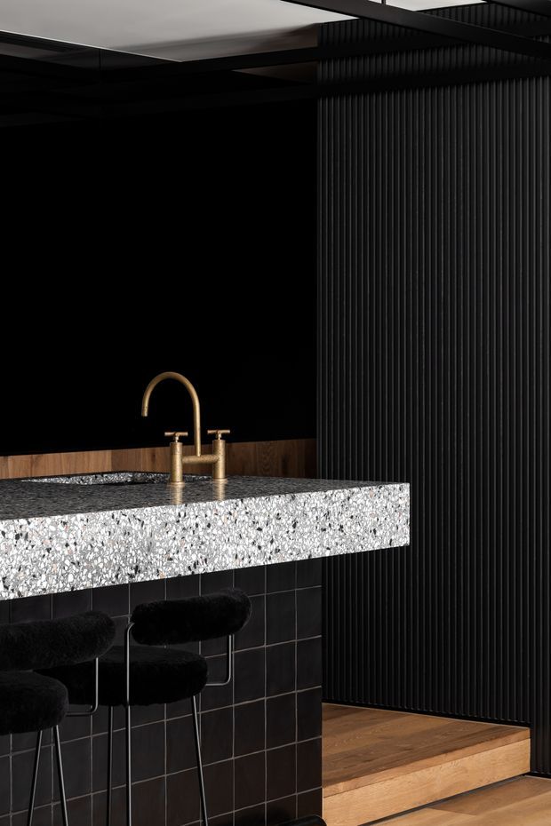 A dramatic, dark-toned contemporary kitchen or home bar featuring a terrazzo island with a thick speckled benchtop, black tiled base, and brushed brass tapware. Black vertical ribbed wall panelling creates a strong textured backdrop, while warm timber flooring and a raised timber step add contrast. Several black upholstered stools sit at the island, and a large sculptural ceiling element is visible above.