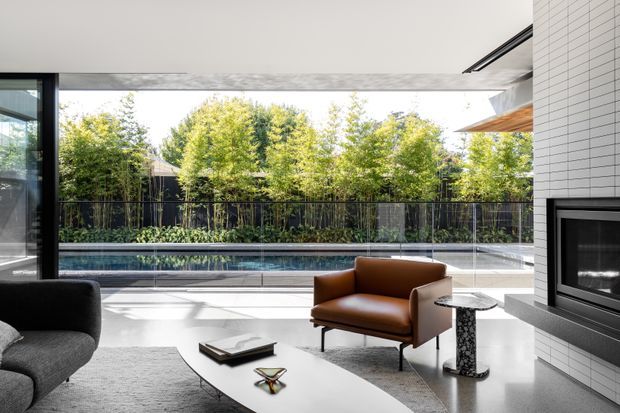 A bright contemporary living room opening onto a pool terrace and landscaped courtyard. The room features floor-to-ceiling glazing, a dark sofa, brown leather armchair, sculptural white coffee table, polished light flooring, and a white tiled fireplace wall with a built-in black fireplace. A large covered outdoor area and swimming pool are visible beyond the glazing.