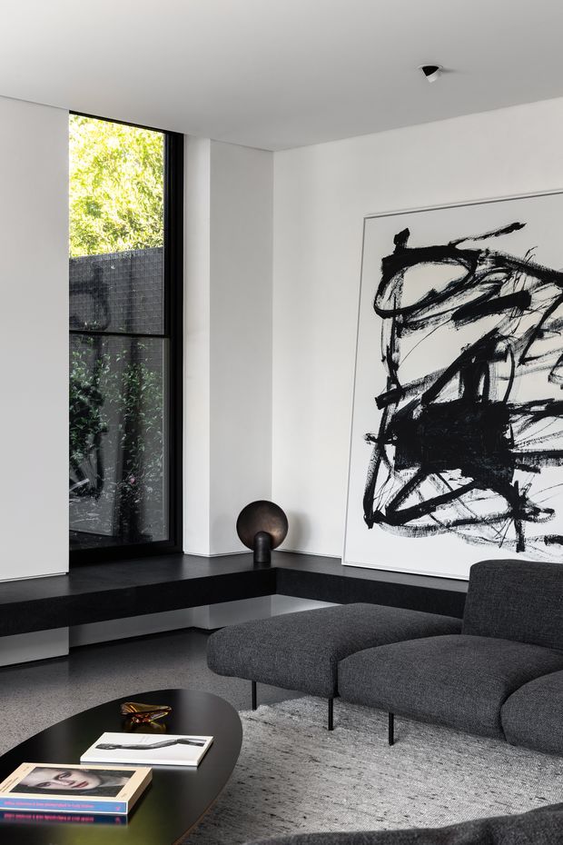 A refined contemporary living room with white walls and ceiling, a large black-framed floor-to-ceiling window overlooking a planted courtyard, and a built-in dark window seat or low bench. A charcoal grey upholstered lounge chair and ottoman sit on a textured grey rug beside a round black coffee table with books and a small decorative object. A large monochrome abstract artwork dominates the wall, complemented by a dark spherical sculpture. The space has a minimalist, monochromatic palette with strong black-and-white contrast and restrained furnishings.