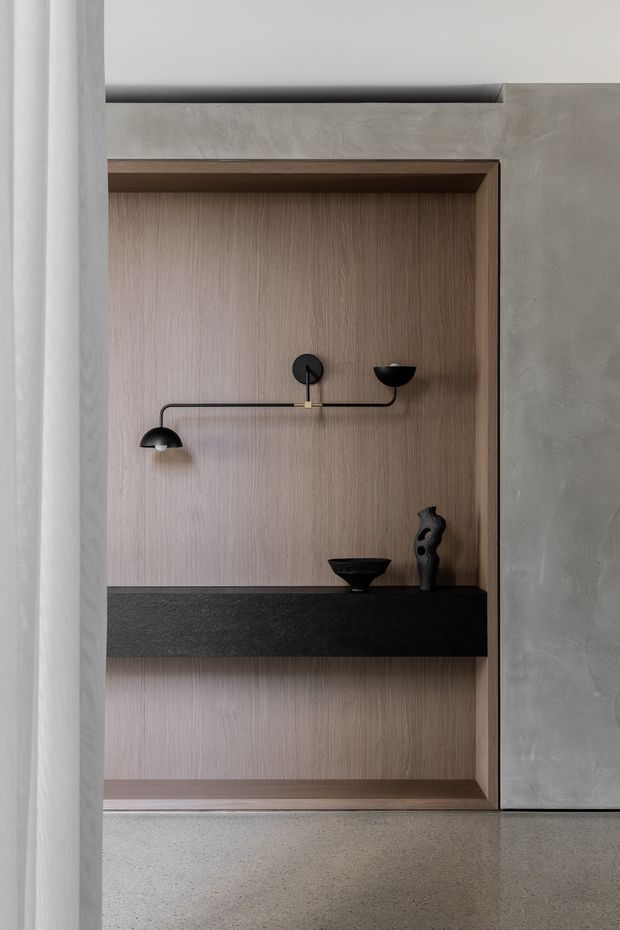 Close-up interior view of a minimalist built-in display niche. Warm vertical-grain timber panels form the recessed back and lower compartment, framed by darker timber trim and a thick black stone or dark composite shelf. A sculptural black wall light and small decorative objects sit within the niche. The surrounding wall has a smooth grey textured finish, with a pale speckled hard floor visible below.