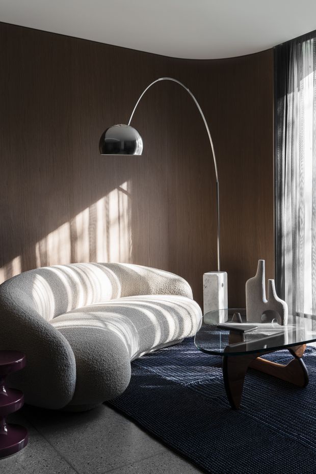 A refined contemporary living room with a sculptural cream boucle sofa, dark curved timber wall panelling, a chrome arched floor lamp, glass coffee table, sculptural neutral decor, and dark textured rug. Floor-to-ceiling sheer curtains bring in strong natural light, creating dramatic shadows across the seating and wall.