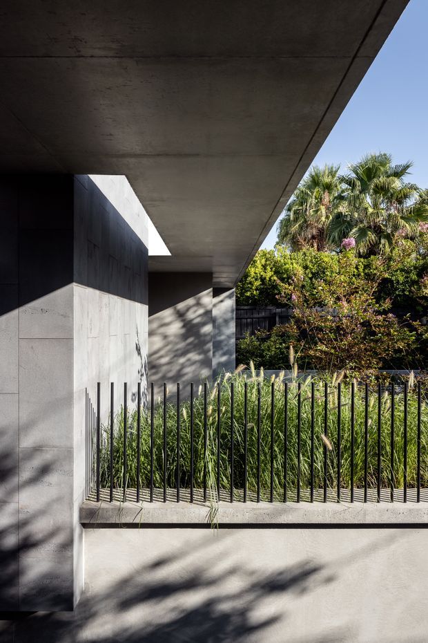 A minimalist modern residential exterior featuring a deep cantilevered concrete roof or canopy, dark and light concrete walls, and a narrow planted courtyard behind a vertical black metal fence. Strong sunlight creates pronounced shadows across the rendered surfaces, with mature palms and greenery visible beyond.