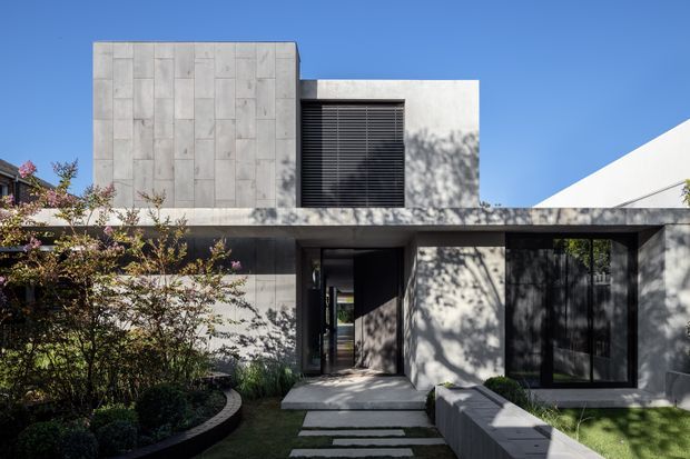 A modern two-storey residential facade with a minimalist geometric composition. The exterior uses large-format pale grey concrete or stone panels, dark-framed glazing, and a prominent black louvred window. A recessed entry is approached by stepping-stone paving through landscaped garden beds, with strong sunlight and tree shadows across the facade.