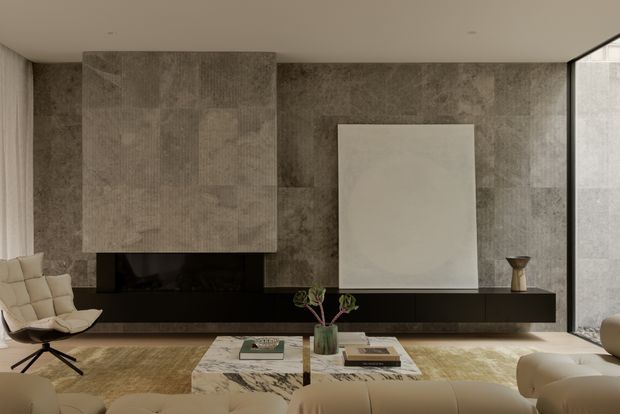 A refined contemporary living room with a long dark built-in media console, a concealed fireplace or TV zone, and a dramatic textured stone feature wall. Large pale artworks, neutral upholstered seating, a marble coffee table, and a floor-to-ceiling corner window create a warm, gallery-like interior.