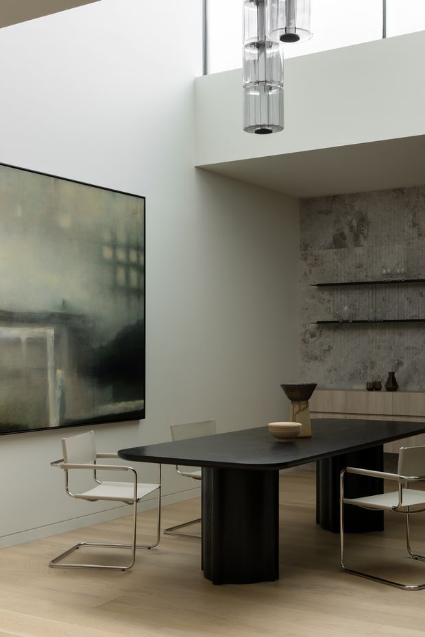 Sophisticated contemporary dining room with a large black oval dining table, sculptural cylindrical pedestal base, and minimalist cantilevered chrome-and-white dining chairs. Pale timber flooring and warm off-white walls contrast with a textured grey feature wall containing slim floating shelves and low built-in cabinetry. A large abstract artwork and clustered cylindrical glass pendant lights add restrained visual interest. The high ceiling and visible upper-level void create an airy, refined atmosphere.