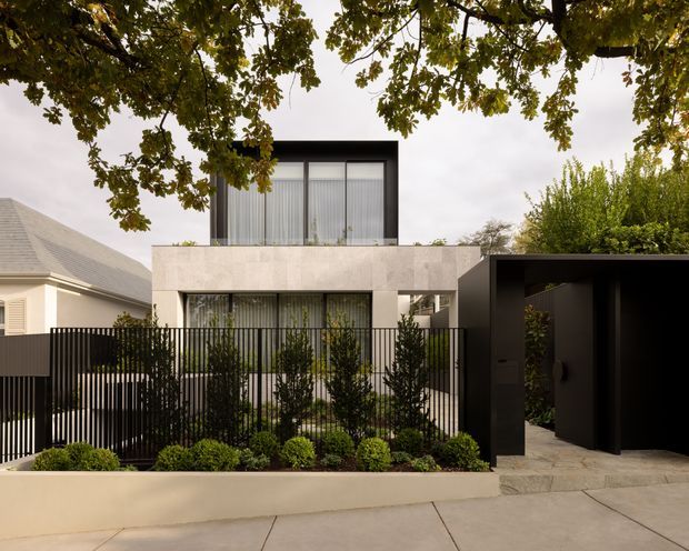 Street-facing view of a contemporary two-storey residence with a pale rendered or stone-like facade, expansive upper-level glazing, dark window frames, and a black slatted front fence. A dark covered entry or carport structure sits to the right, with landscaped planting and a paved frontage in the foreground. Mature tree branches frame the image, while a neighbouring traditional pitched-roof house is partially visible to the left.