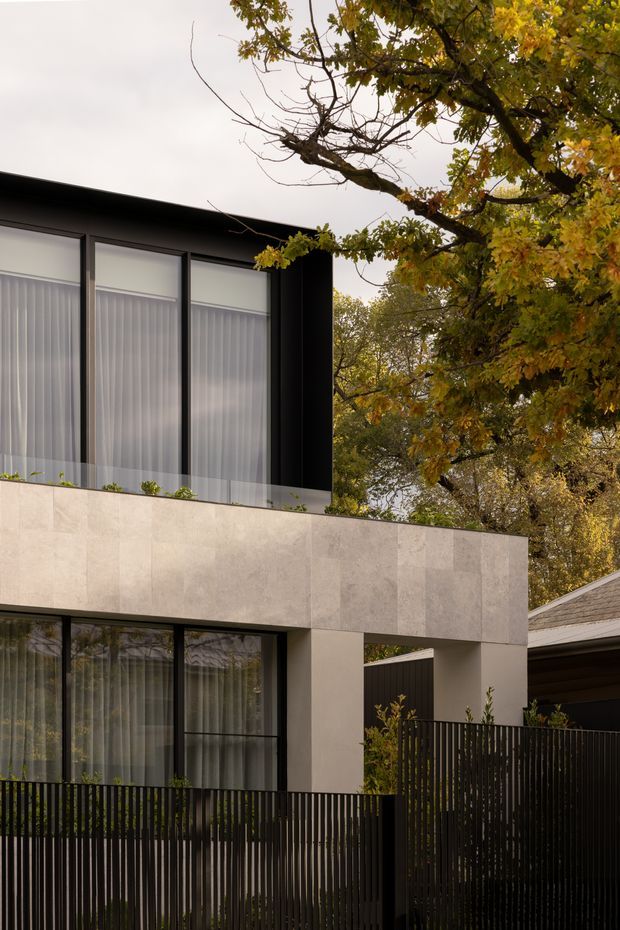 A modern two-storey residential facade photographed from outdoors, featuring large floor-to-ceiling windows with sheer curtains, dark metal framing, a pale stone or rendered upper-level band, and a black vertical-slat front fence. Mature autumn trees surround the building, while part of an adjacent pitched-roof structure is visible at right.