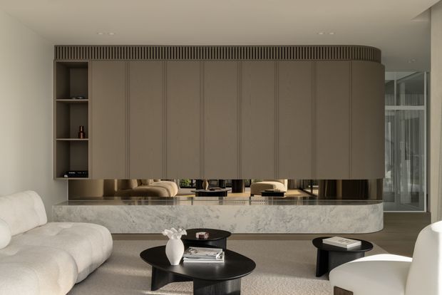 A refined contemporary living room centred on a sculptural curved entertainment or storage wall with fluted detailing, open shelving, and integrated reflective niches. A pale marble-clad low cabinet or console runs beneath it, paired with a cream boucle sectional, sculptural lounge chairs, and low black round coffee tables. The space has soft neutral tones, restrained lighting, and glazing at the right side.