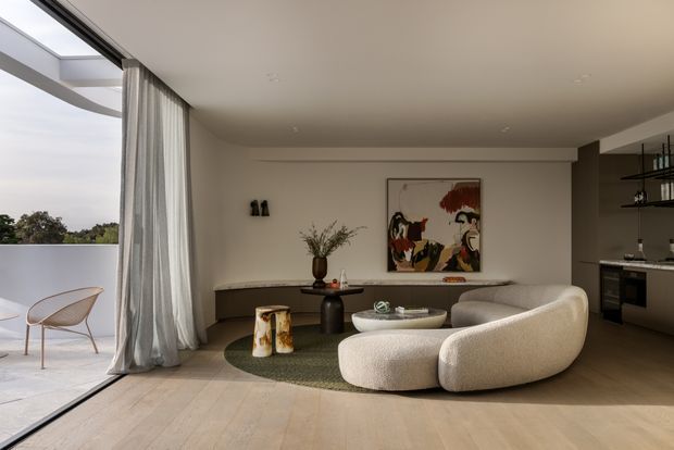 A refined open-plan living room with a sculptural curved cream sofa and matching rounded lounge chair arranged around a low circular coffee table and textured green rug. Full-height glazed sliding doors open to a terrace, with sheer curtains, pale timber flooring, a dark built-in console, abstract artwork, and a compact kitchen with dark cabinetry and open shelving visible at the rear. The space has soft neutral tones, restrained contemporary detailing, and abundant natural light.