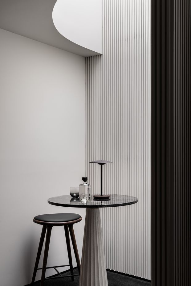 A refined monochromatic interior vignette featuring a small round pedestal table with a dark veined stone top, a sculptural fluted light-toned base, a dark timber or black stool, and a slim table lamp with glassware. The space is defined by curved ceiling geometry, a white wall, a tall vertically ribbed white feature surface, and contrasting dark vertical panelling. The composition is minimalist, dramatic, and highly textural.