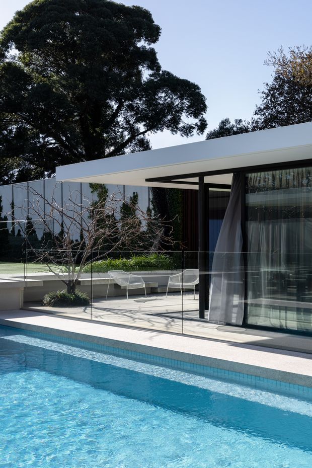 A sleek modern residential pool courtyard with a long turquoise swimming pool in the foreground, pale paving, frameless glass balustrades, and a low flat-roofed pavilion or house. Black-framed floor-to-ceiling glazing opens onto a minimalist terrace furnished with white chairs and a built-in bench. Mature trees and sculptural leafless planting surround the private outdoor space.