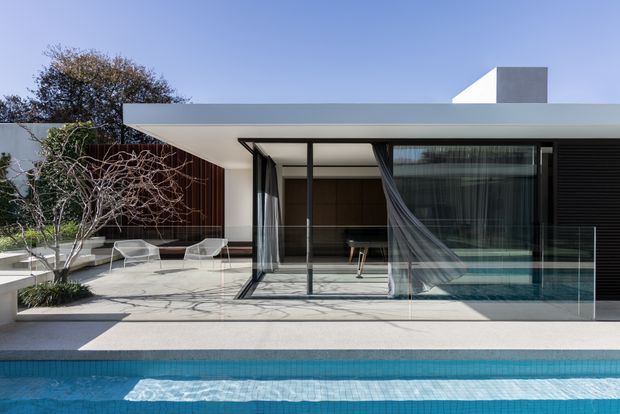 A minimalist modern single-storey residence viewed from the pool, with a broad flat roof, extensive floor-to-ceiling glazing, dark framed sliding doors, and a covered outdoor terrace. The restrained white, grey and dark timber facade opens onto a pale paved patio with sculptural outdoor loungers and a swimming pool in the foreground.