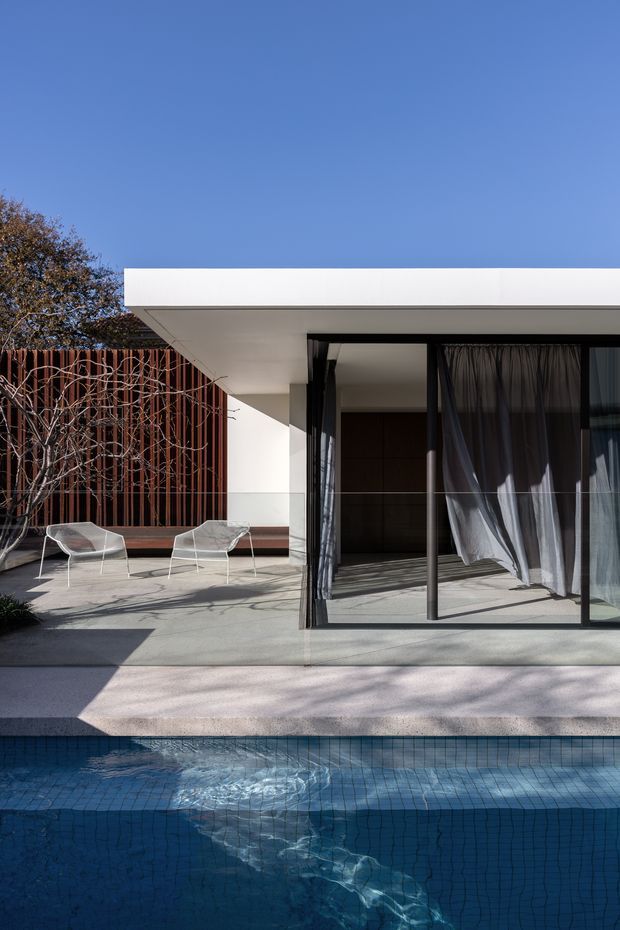 A contemporary residential exterior courtyard viewed from beside a swimming pool. The scene features a crisp flat-roofed white pavilion, expansive dark-framed glazed sliding doors with sheer curtains, a concrete terrace, a rust-toned vertical slatted feature wall, and minimalist outdoor lounge chairs. Bright clear daylight and strong geometric lines create a refined indoor-outdoor connection.