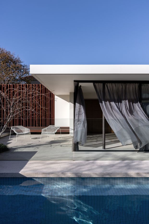 Exterior view of a contemporary flat-roofed residence beside a swimming pool. The architecture features a crisp white projecting roof canopy, extensive dark-framed glazing with sheer curtains, a vertical timber-clad privacy wall, and a pale concrete terrace with outdoor lounge chairs. Strong sunlight and deep shadows create a clean, refined indoor-outdoor setting.