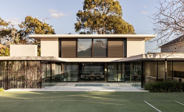 A contemporary two-storey residential building viewed from a landscaped tennis court. The house has a broad flat roof with deep overhangs, a restrained pale rendered facade, extensive floor-to-ceiling glazing, dark timber or dark-finished window shutters, and a glazed central courtyard or living area with a small rectangular pool. Mature trees and adjacent residential structures surround the property.