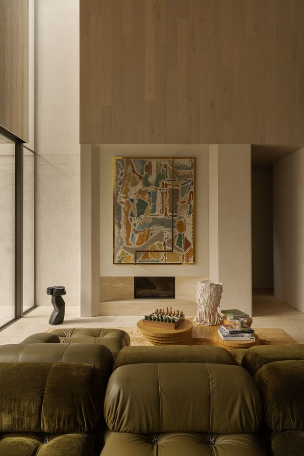 A warm, refined living room viewed from behind a large deep olive-green sectional sofa. The room features a tall natural timber wall and ceiling volume, pale stone or plaster walls, expansive floor-to-ceiling glazing at left, a built-in fireplace, abstract artwork, a round timber coffee table, and sculptural decorative objects. The composition has a softly lit, earthy and luxurious contemporary character.